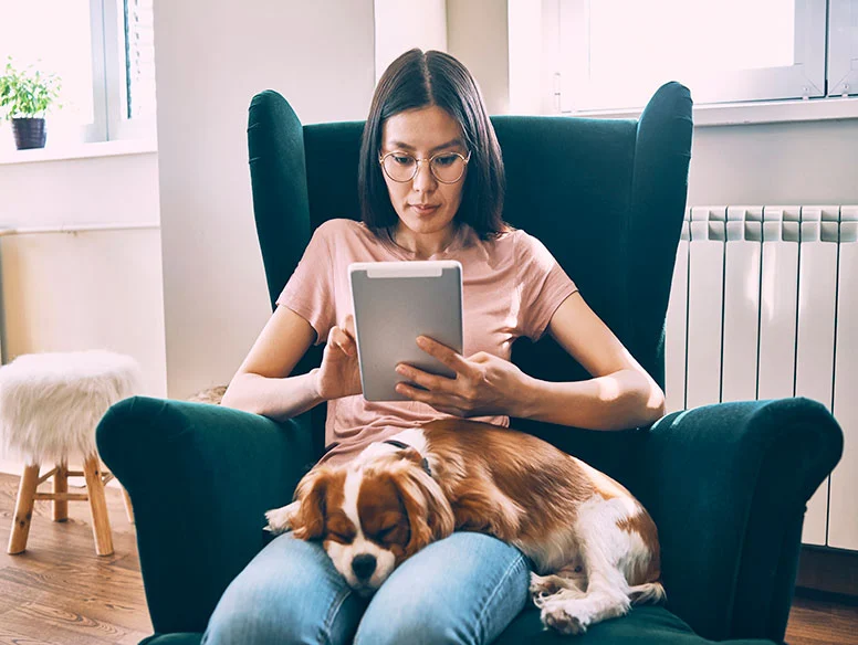 woman sitting down in a chair using a tablet with a dog on her lap.