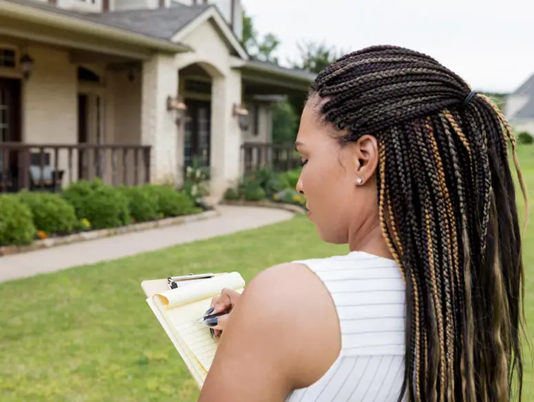 woman in front of a house taking notes on a clipboard.