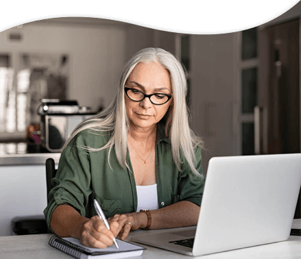 Woman writing on notebook with laptop open at home desk.