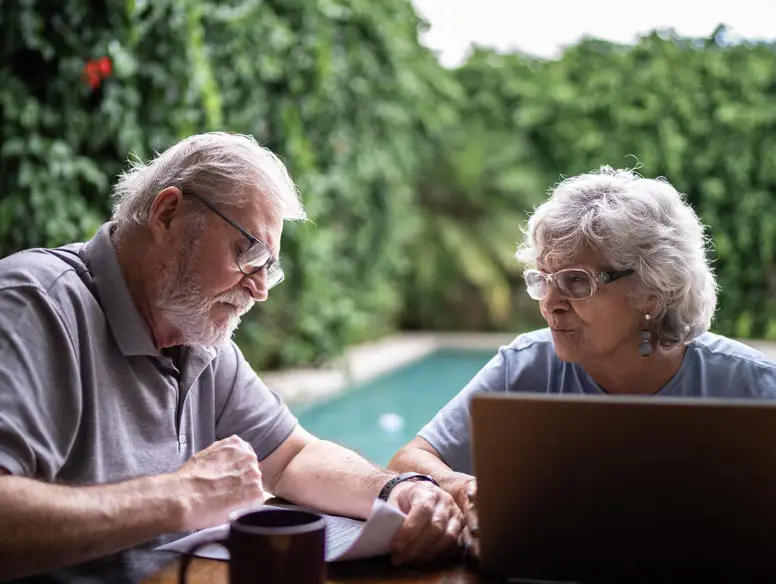 elderly couple sitting at a table, by a pool, while reading documents and using a laptop.