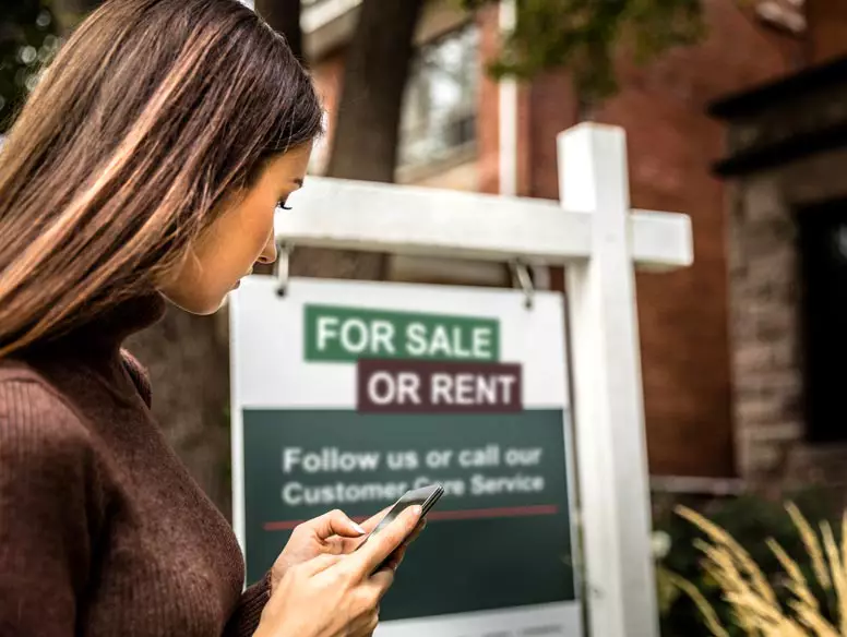 woman checking phone in front of a "sale or for rent" sign.