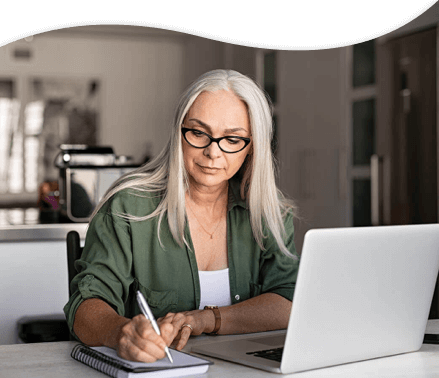 A woman writing in a notebook with a laptop opened.